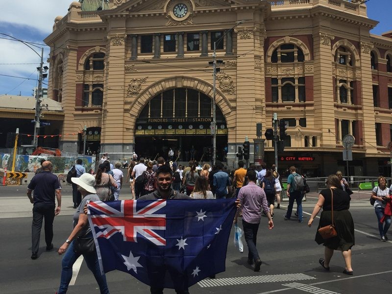 chico con la bandera de Australia en medio de la gente y enfrente de un monumento en la ciudad de Melbourne